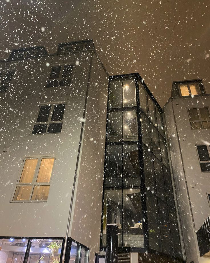 snow falling from the sky in front of two tall buildings with windows and balconies