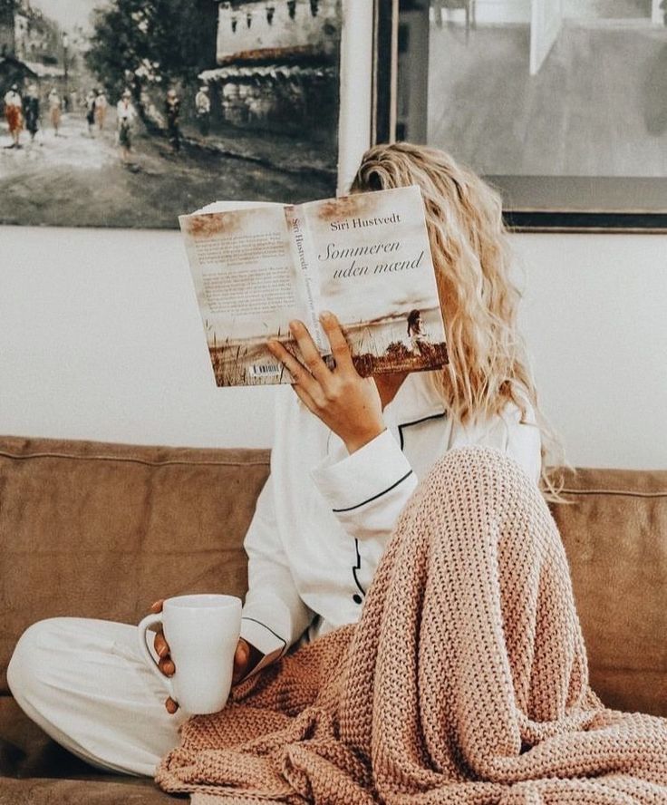 a woman sitting on a couch reading a book and holding a coffee cup in her hand