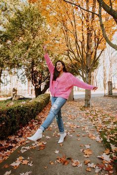 a woman in pink jacket and jeans dancing on sidewalk next to trees with yellow leaves