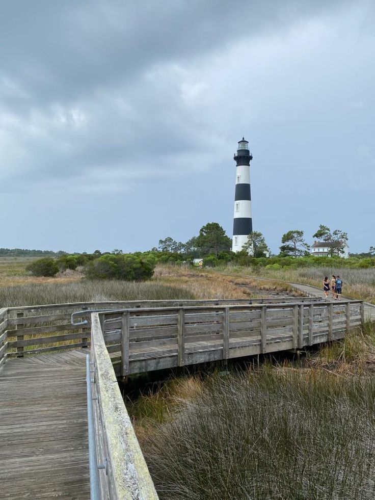 people are walking on a boardwalk near a light house in the distance with tall grass