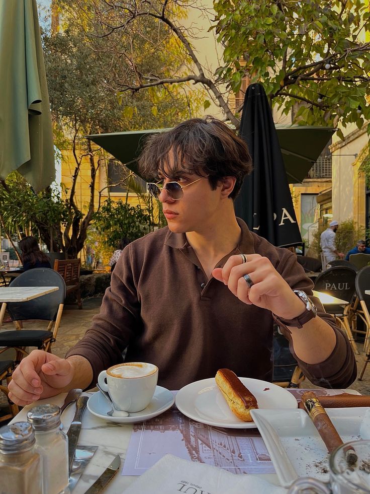 a man sitting at an outdoor table with food in front of him and drinking coffee