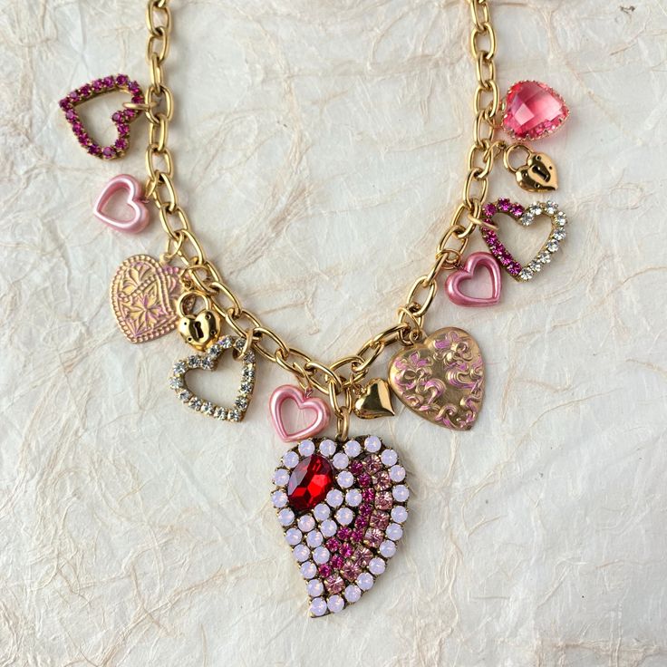 "The sweetest Valentine's Day charm necklace filled with an alluring mix of dangling heart charms. Heart charms include classic brass heart charms, some with shimmering Swarovski rhinestones, playful little pink hearts, and a bold rhinestone collage heart focal point to steal the show. These heart charms wire wrapped to perfection dangle from a high-quality gold-plated cable chain. This necklace has a solid and nice heavy weight to it for durability. 18\"l with 3\" extension. Lobster claw closure. Handmade in USA. ~NOTE: Some of the small heart charms are upcycled vintage pieces with limited quantity in our supply so they may vary from the photo but we are confident you'll love them!" Pink Heart Pendant Charm Necklace With Lobster Clasp, Pink Heart Charm Necklace With Heart Pendant, Pink Heart-shaped Charm Necklaces With Lobster Clasp, Pink Heart-shaped Charm Necklace With Lobster Clasp, Pink Metal Heart Necklace For Valentine's Day, Pink Heart Charm Necklaces For Jewelry Making, Pink Pendant Charm Necklace With Dangling Charms, Pink Pendant Charm Necklaces With Dangling Charms, Pink Heart Pendant Jewelry With Rhinestones