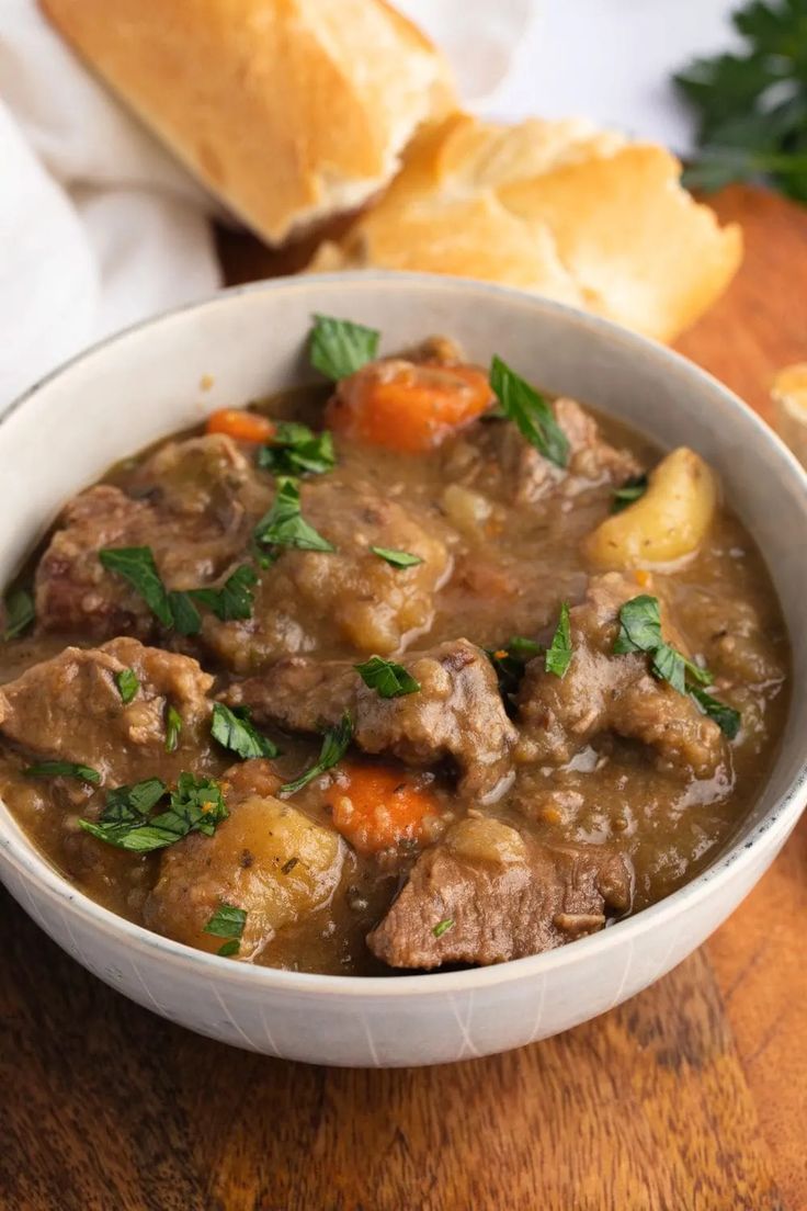 a white bowl filled with meat and vegetables on top of a wooden table next to bread