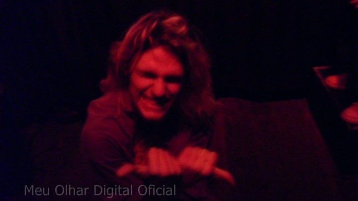 a man with long hair sitting on a couch in front of a purple light and pointing at something