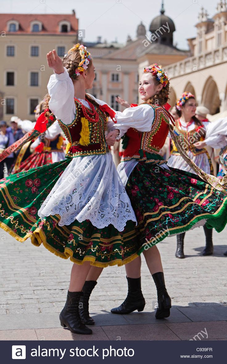 two women dressed in folk costumes dancing on the street while others watch from behind them