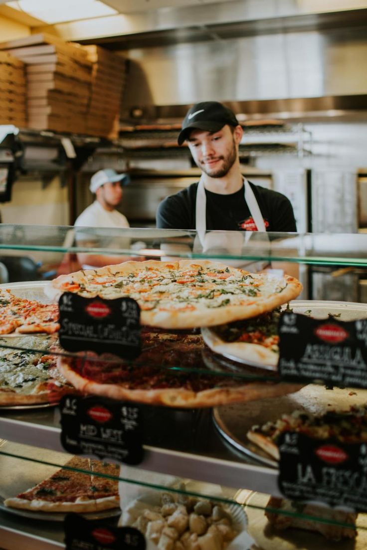 a man standing behind a display case filled with pizzas