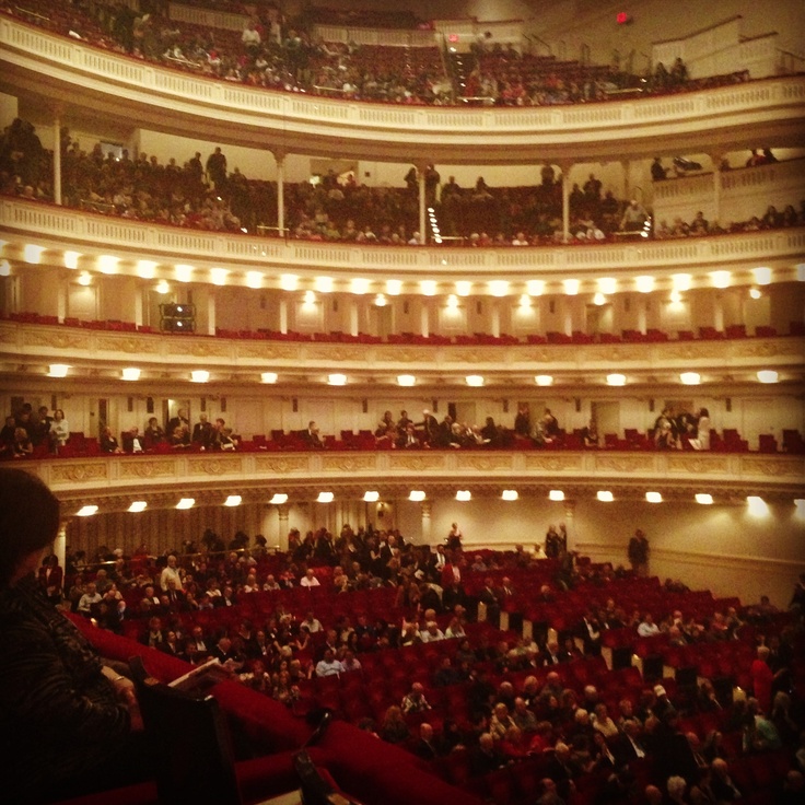 an auditorium filled with lots of people sitting in red seats and looking up at the ceiling