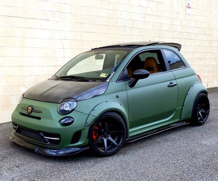 a small green car parked in front of a white building with black rims and red brake pads