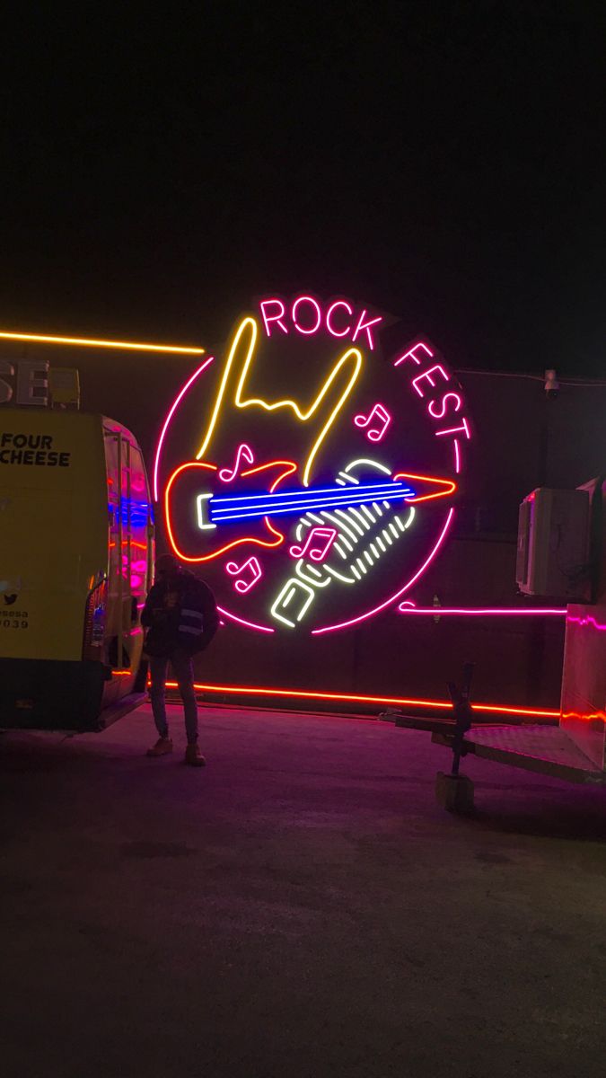 a neon sign that says rock fest with a guitar on it's side and people standing in front of it