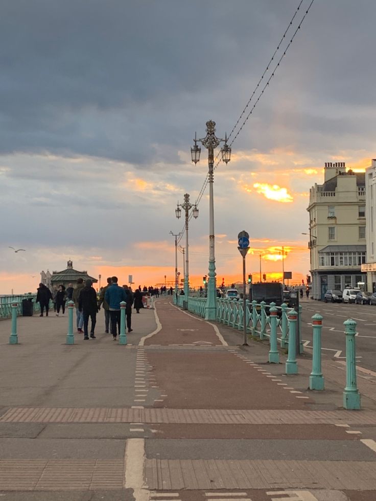 people are walking on the sidewalk near some buildings and street lights as the sun sets