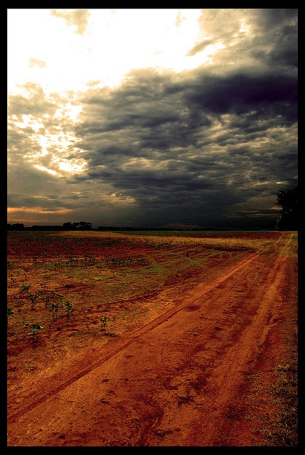 a dirt road in the middle of an open field under a dark sky with clouds