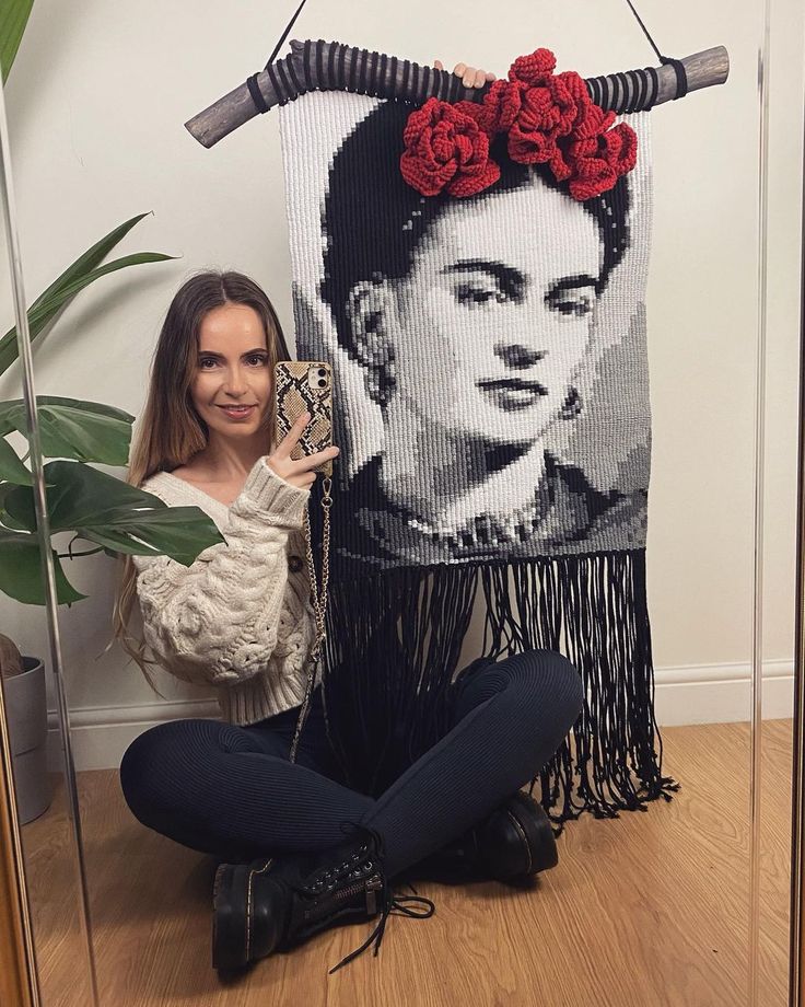 a woman sitting on the floor next to a wall hanging with a portrait of fridace