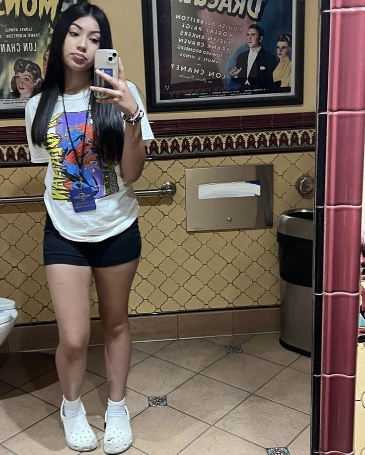 a young woman taking a selfie in a bathroom with posters on the wall behind her