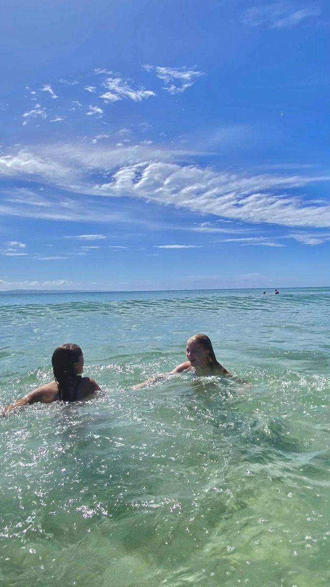 two people swimming in the ocean under a blue sky with wispy white clouds