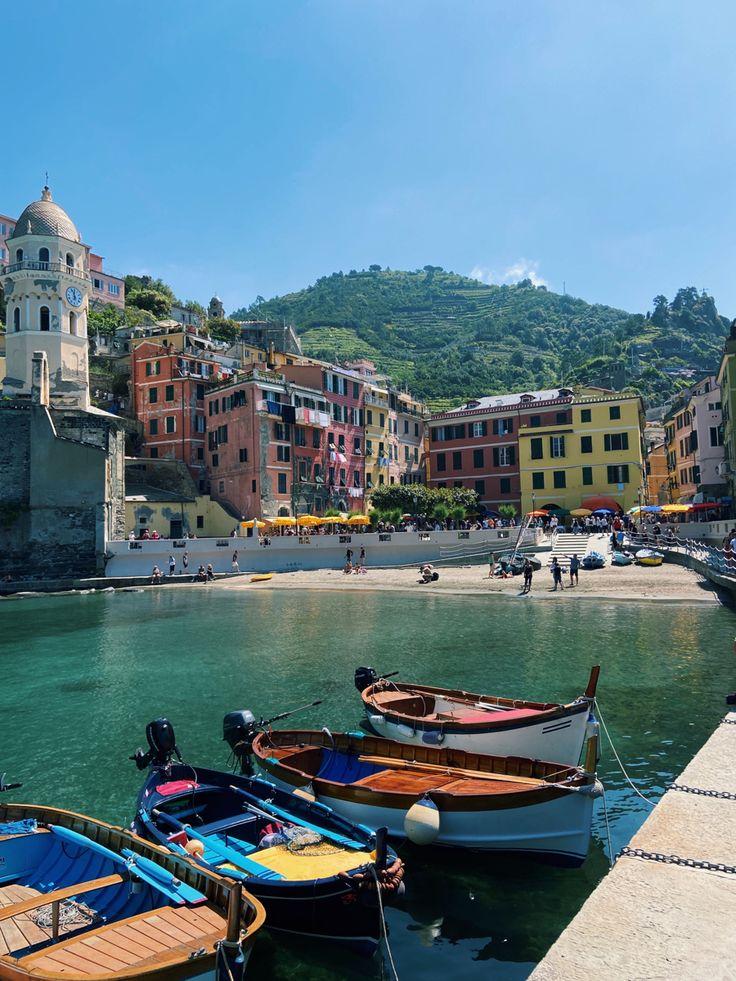 several small boats tied to the dock in front of some colorful buildings and water with people walking around them