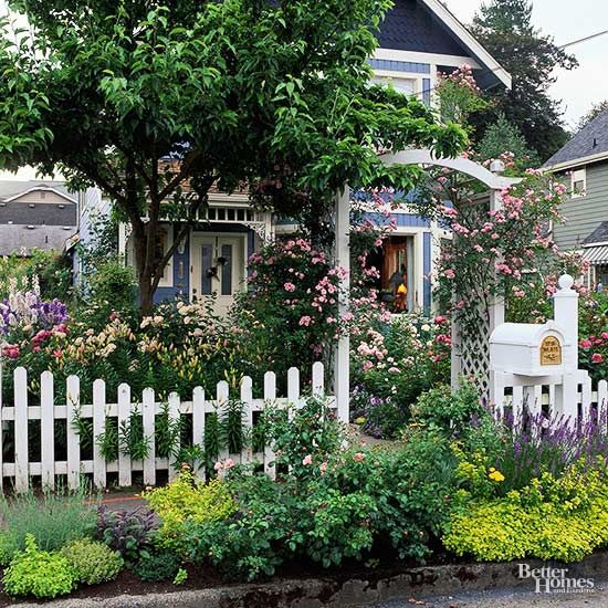 a white picket fence in front of a house with lots of flowers and trees around it