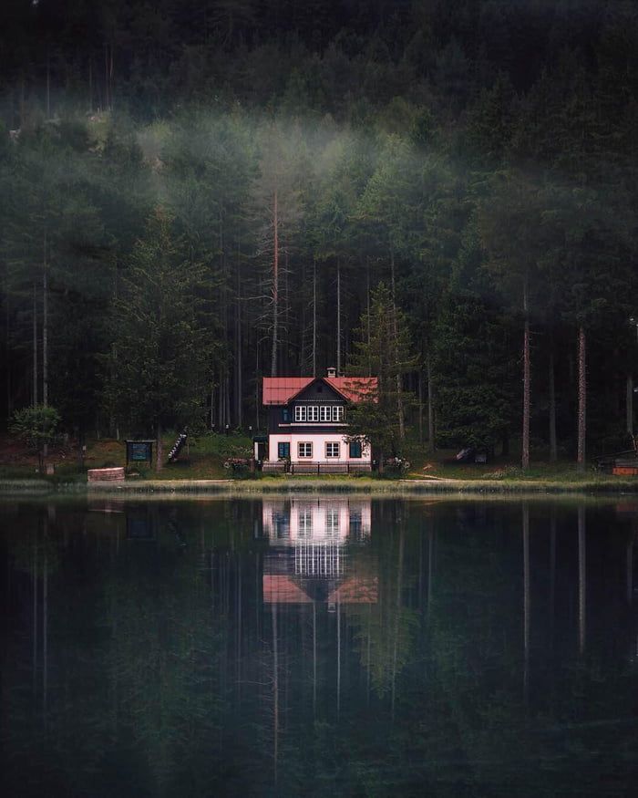 a house sitting on the shore of a lake surrounded by trees with fog in the air