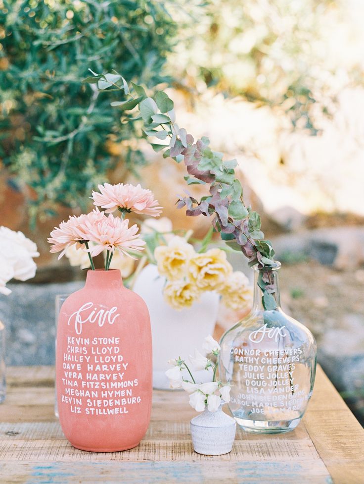 two vases with flowers on top of a wooden table in front of a bush