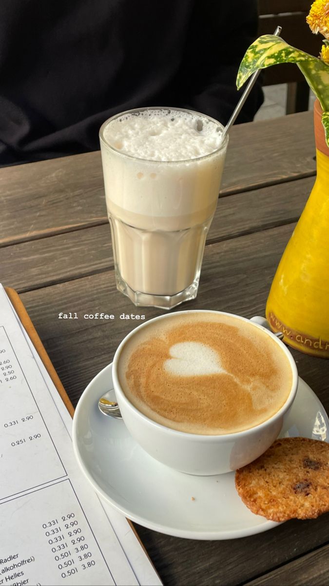 a cappuccino and cookie sit on a table next to a yellow vase with flowers