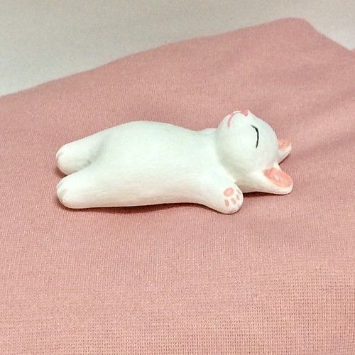 a white cat figurine laying on top of a pink bed sheet with its eyes closed