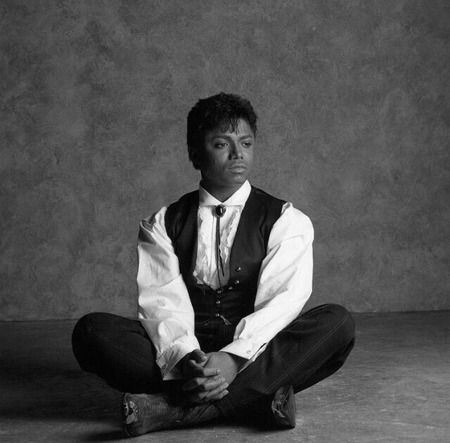 a man sitting in the middle of a floor with his legs crossed, wearing a vest and tie