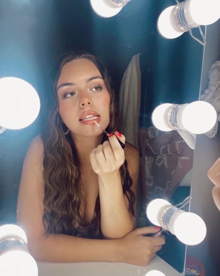 a woman sitting in front of a mirror with lights on her face and holding a lipstick