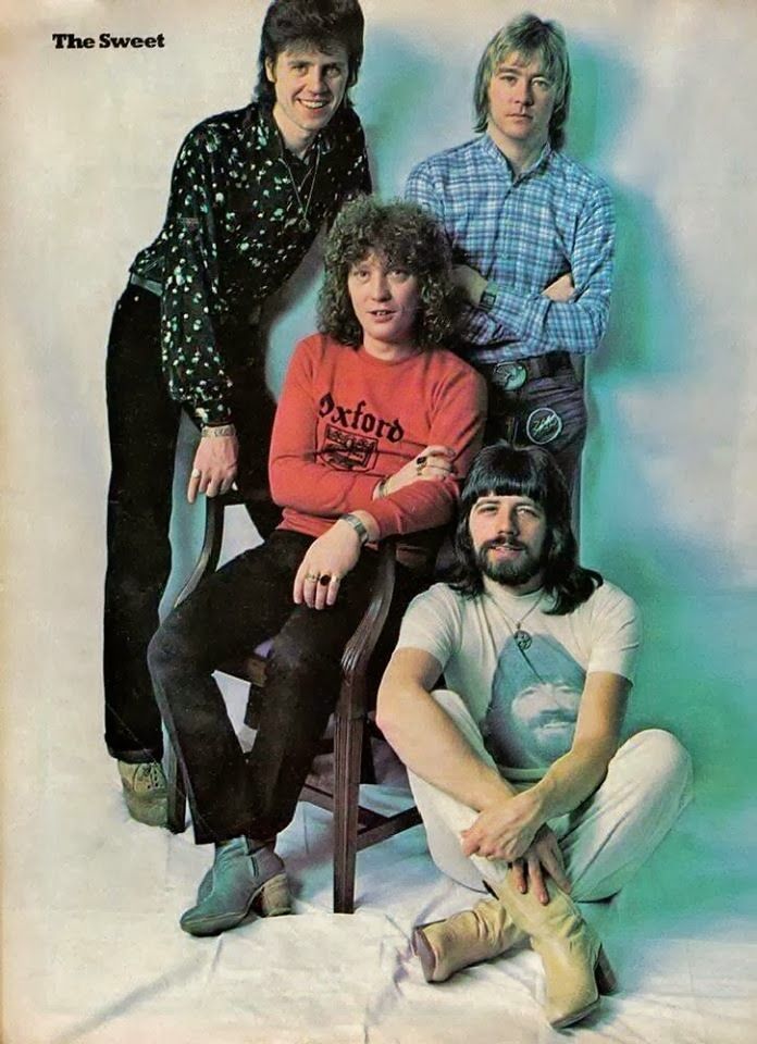 the bee gees are posing for a photo in front of a blue background, with one man sitting on a chair