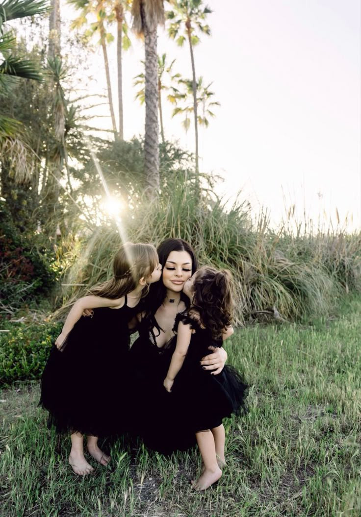 three women in black dresses hugging each other on the grass with palm trees behind them