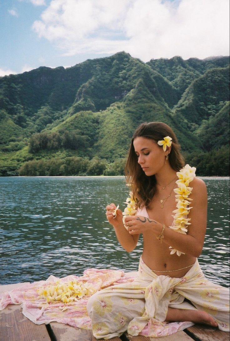 a woman sitting on top of a wooden pier next to a body of water with flowers in her hair