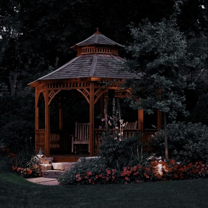 a wooden gazebo surrounded by flowers and greenery in the evening time with lights on