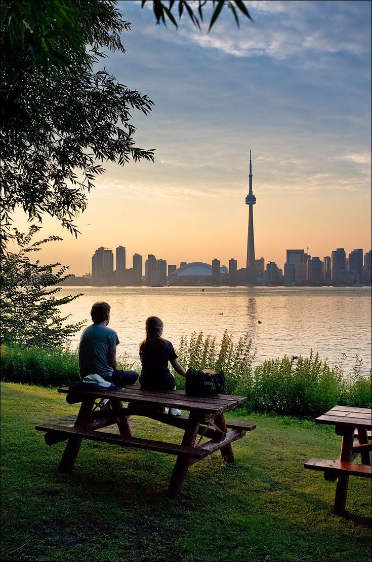 two people sitting on picnic benches in front of a lake with the city skyline in the background