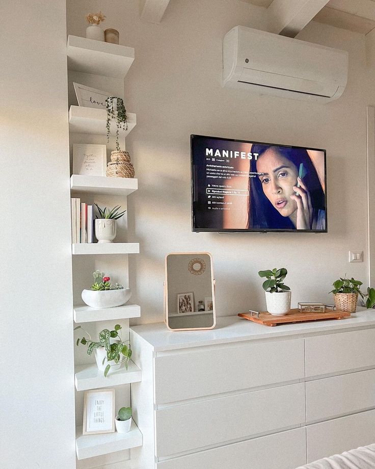 a flat screen tv mounted on the wall above a white dresser with books and plants