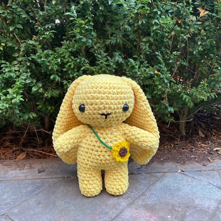 a crocheted yellow stuffed animal holding a sunflower in front of some bushes