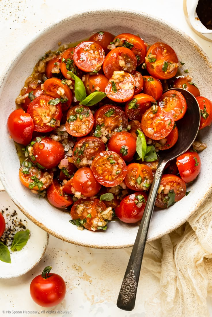 a white bowl filled with tomatoes and basil on top of a table next to a spoon