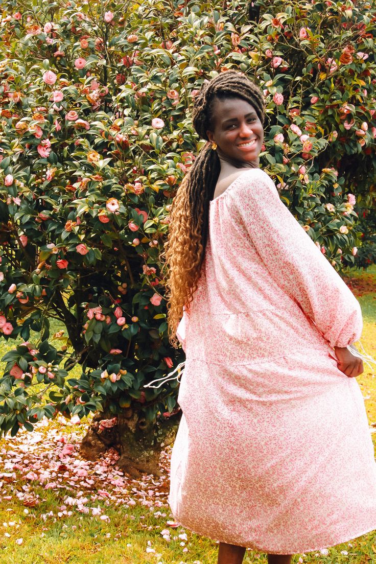 a woman in a pink dress standing next to a bush full of flowers and trees