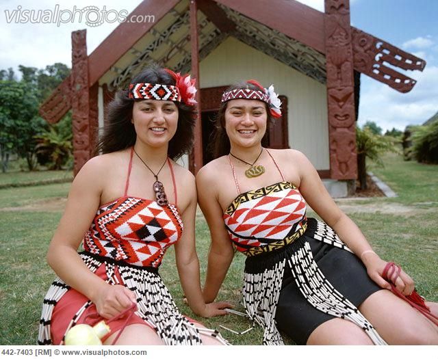 two women dressed in native clothing sitting on the grass near a small building with a wooden structure behind them