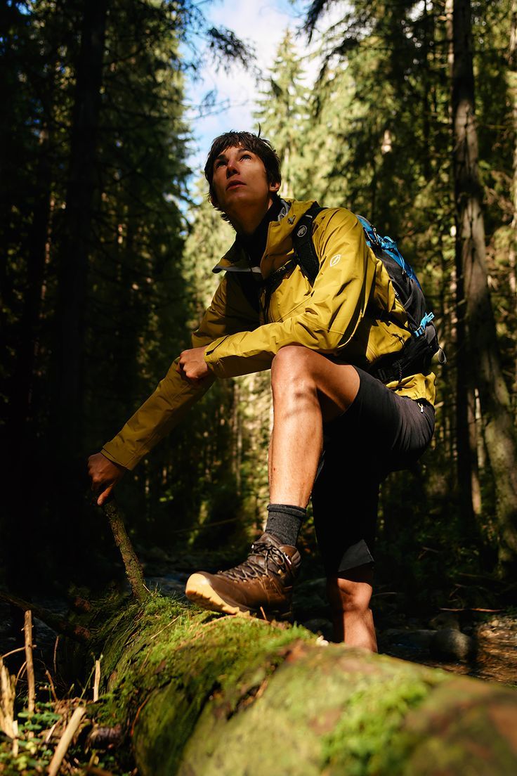 a man in yellow jacket and black shorts standing on tree stump with backpack over his shoulder