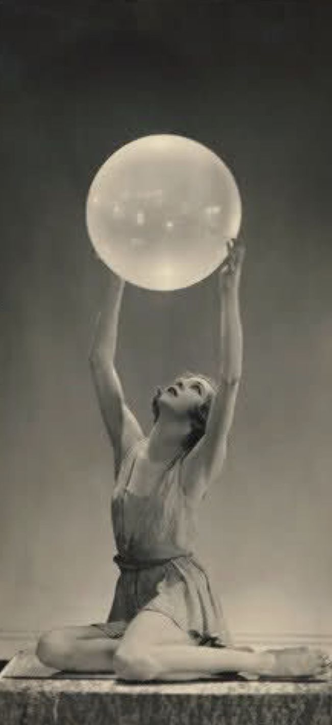 a woman sitting on the ground holding up a large white ball above her head, in front of a black and white background