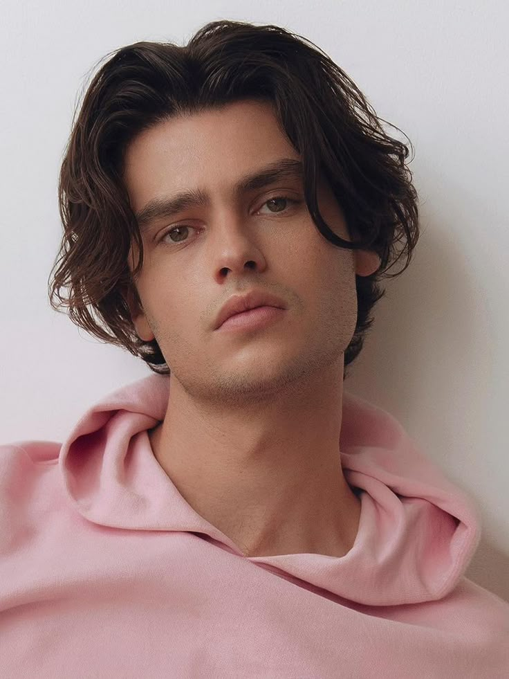 a man with long hair wearing a pink hoodie looking at the camera while leaning against a wall