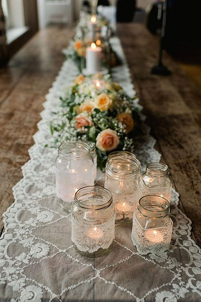 a long table is set with candles and flowers on the table runner, along with glass jars filled with water