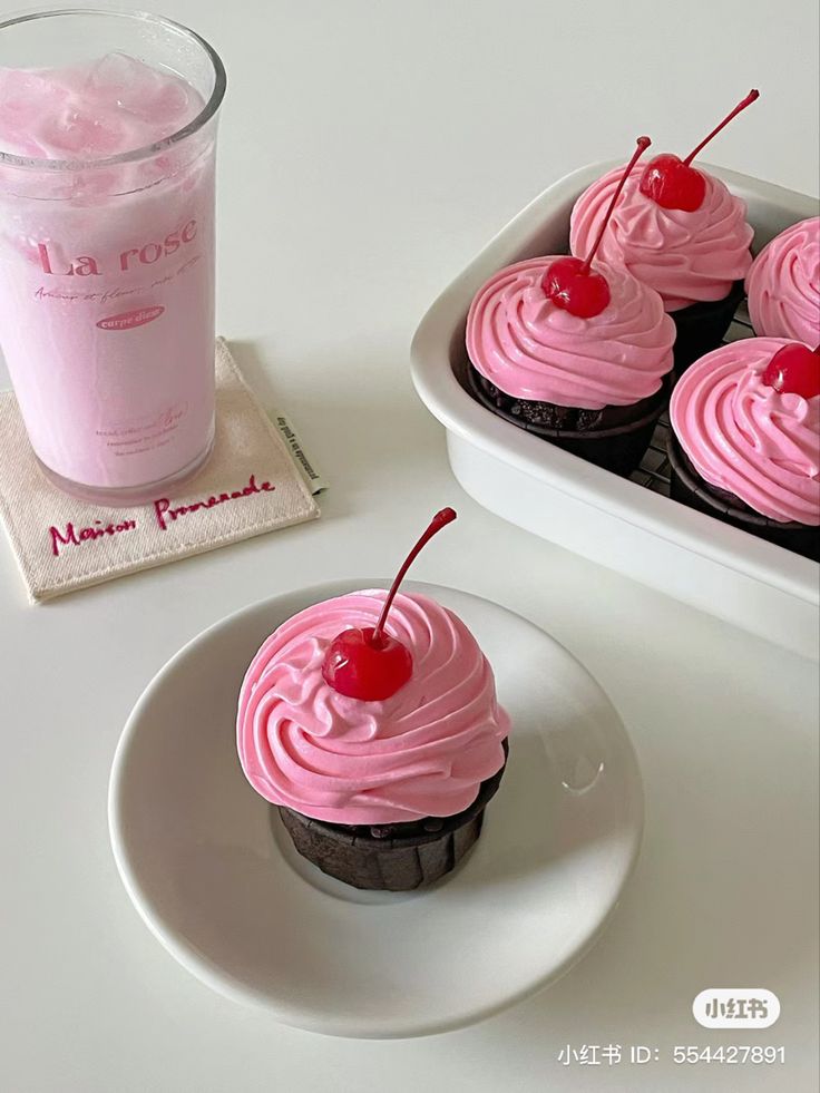 two cupcakes with pink frosting and cherries are on a plate next to a drink