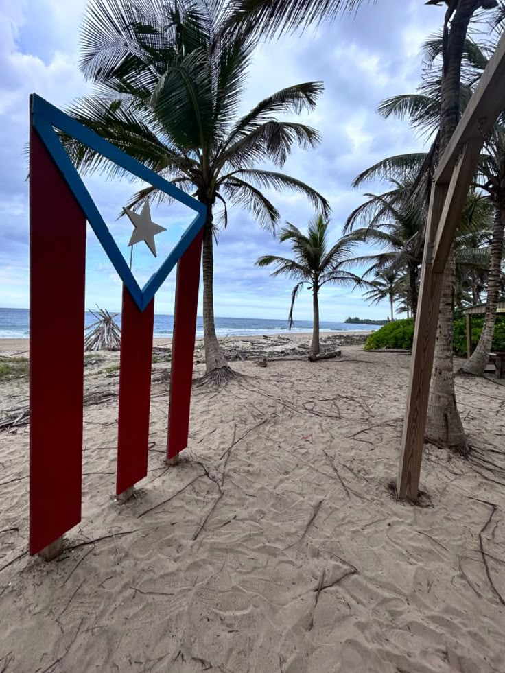 a red, white and blue sign sitting on top of a sandy beach next to palm trees