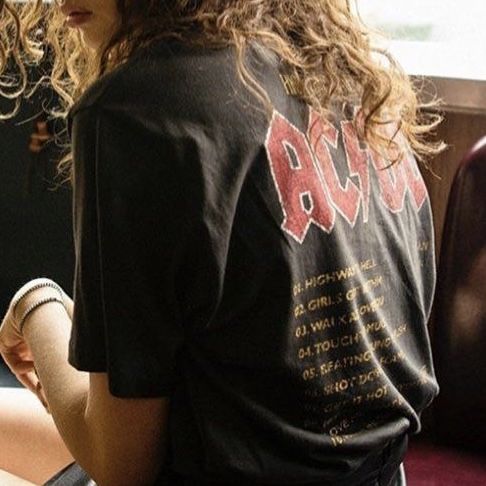 a woman with long curly hair sitting on a bus looking at her cell phone while wearing a t - shirt that says aca