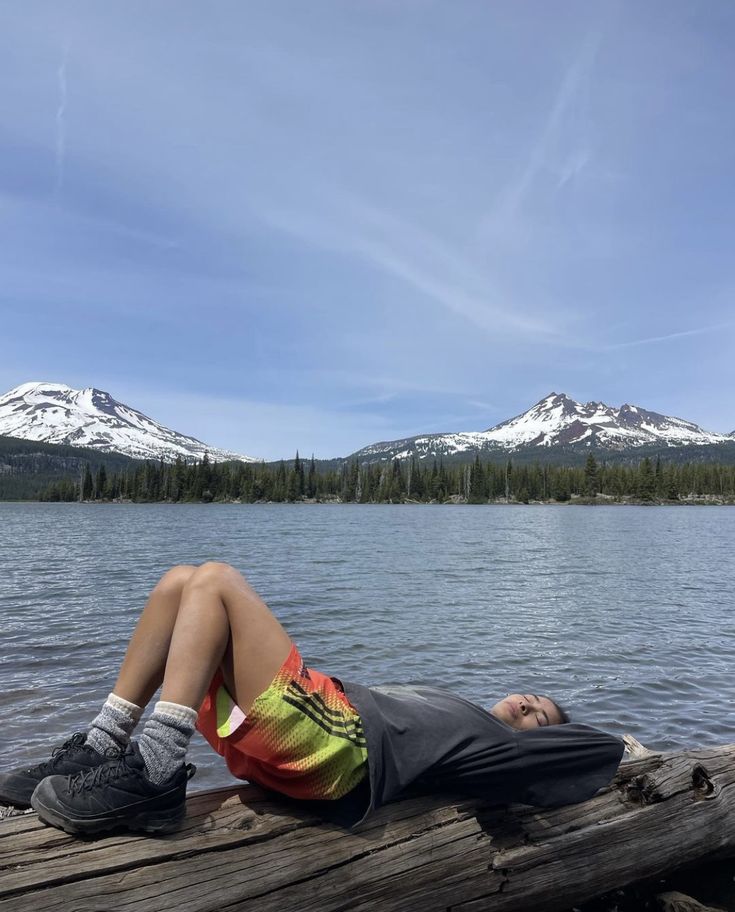 a man laying on top of a wooden dock next to a lake with snow covered mountains in the background