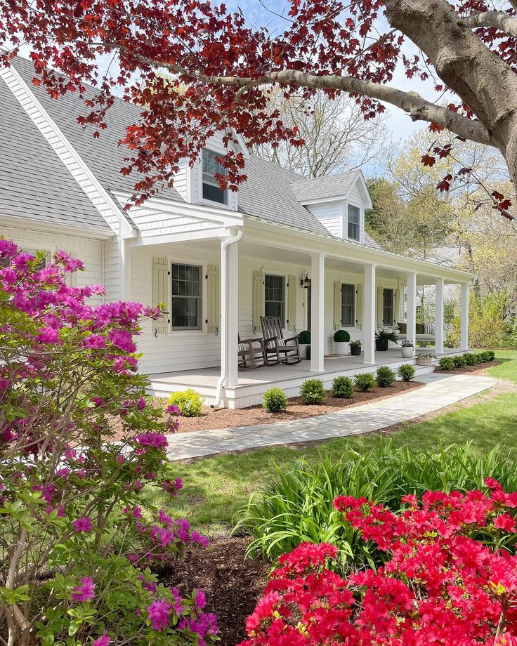 a white house with red flowers in the foreground and trees on either side of it