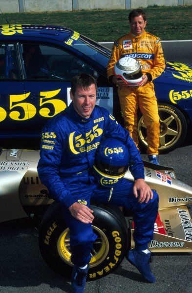 two men in racing suits sitting on the ground next to a race car and another man standing behind them