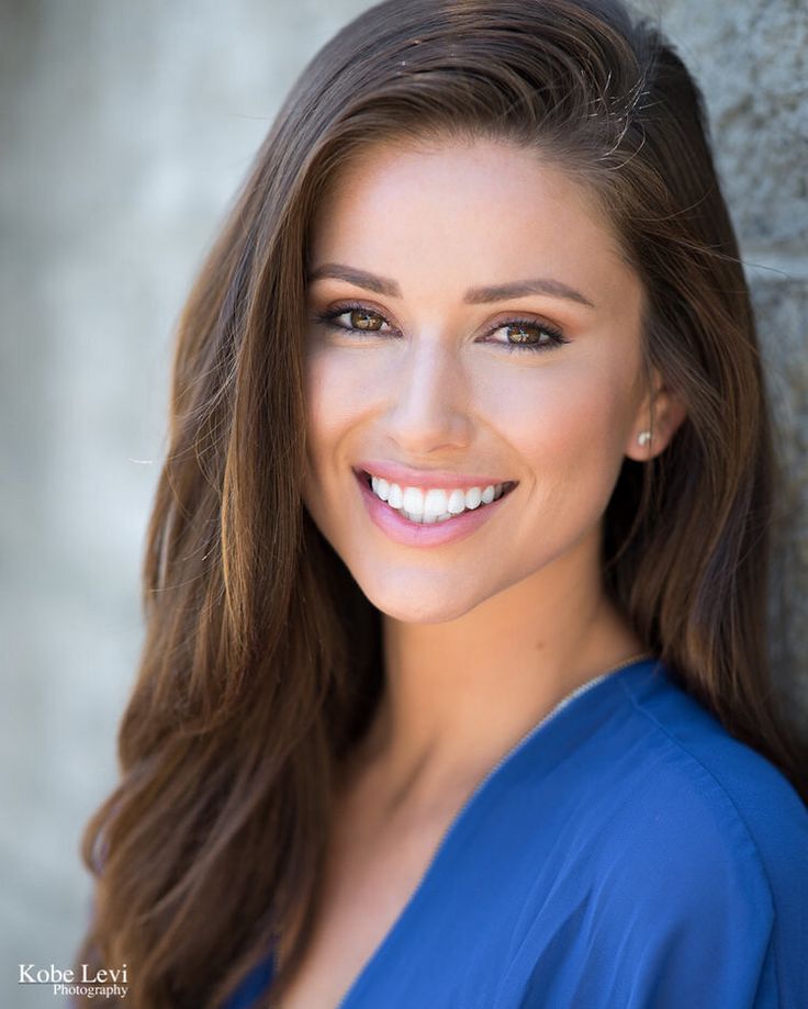 a woman with long brown hair and blue dress smiling at the camera while leaning against a stone wall