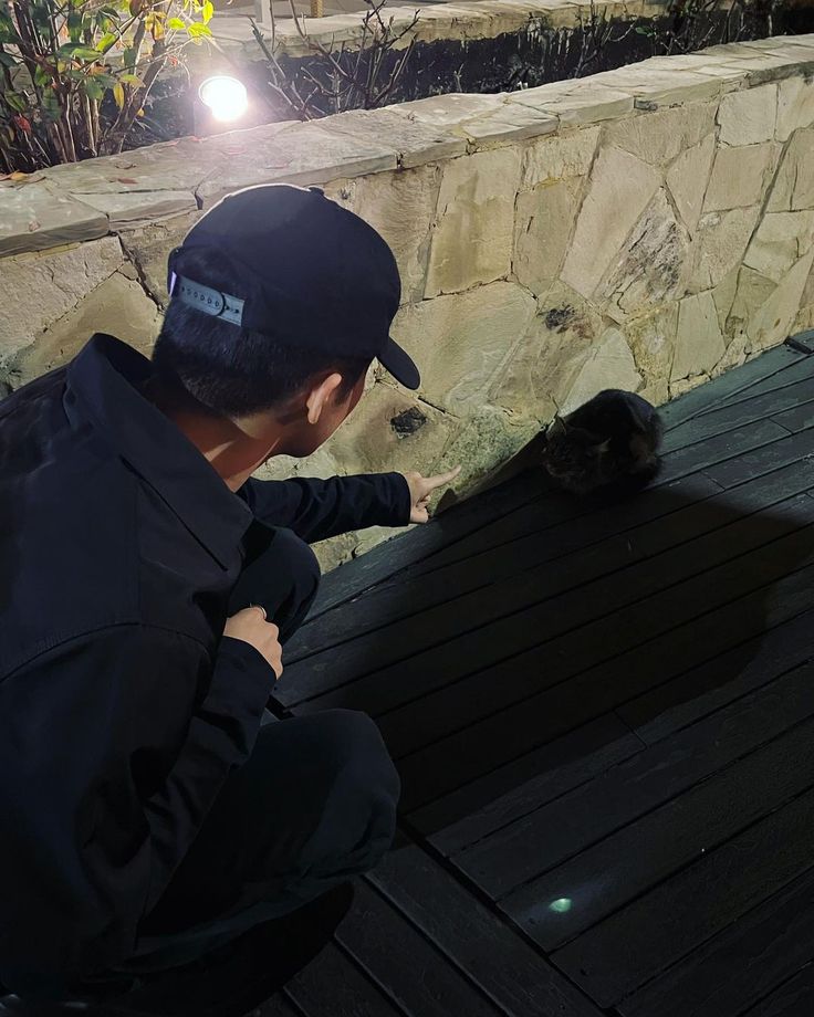 a man kneeling down next to a cat on top of a wooden floor in front of a stone wall