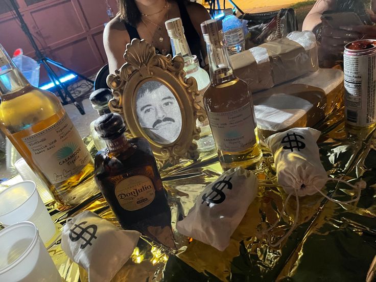 a woman sitting at a table with bottles of liquor in front of her and a mirror