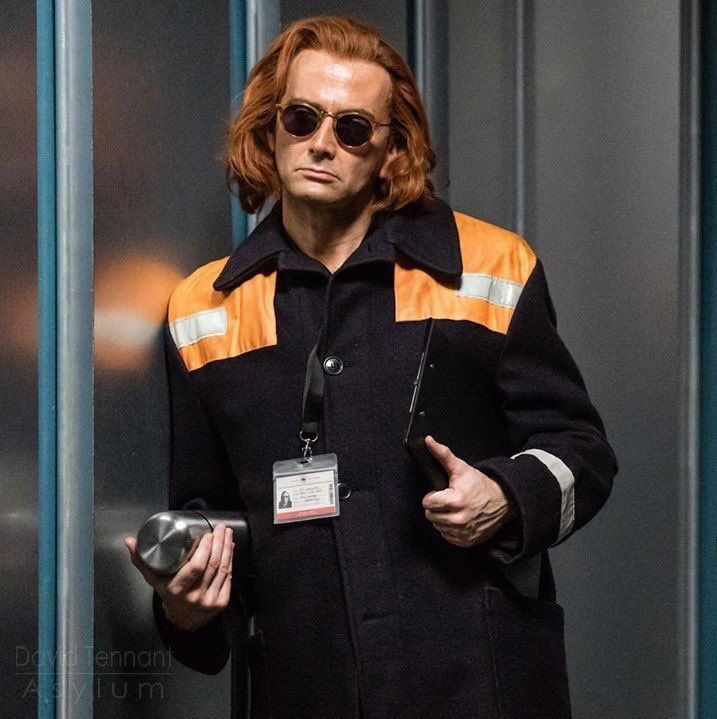 a man with red hair and sunglasses standing in front of a glass door wearing a black coat